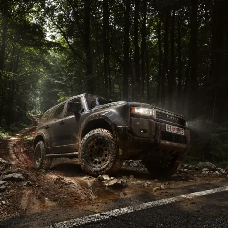 Toyota Land Cruiser with General Grabber AT3 Plus tyres crossing a muddy, rocky forest track beside a road.