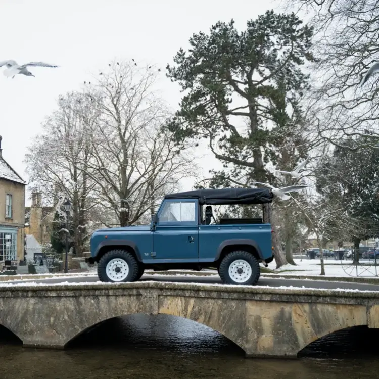 Blue Land Rover Defender with white steel wheels on a snow-covered stone bridge.