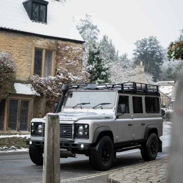 Silver Land Rover Defender with black wheels and a roof rack on a village street beside snow-covered cottages.