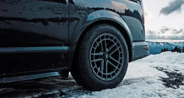 Close-up of a grey alloy wheel on a dark campervan parked on snow, with mountains in the background.